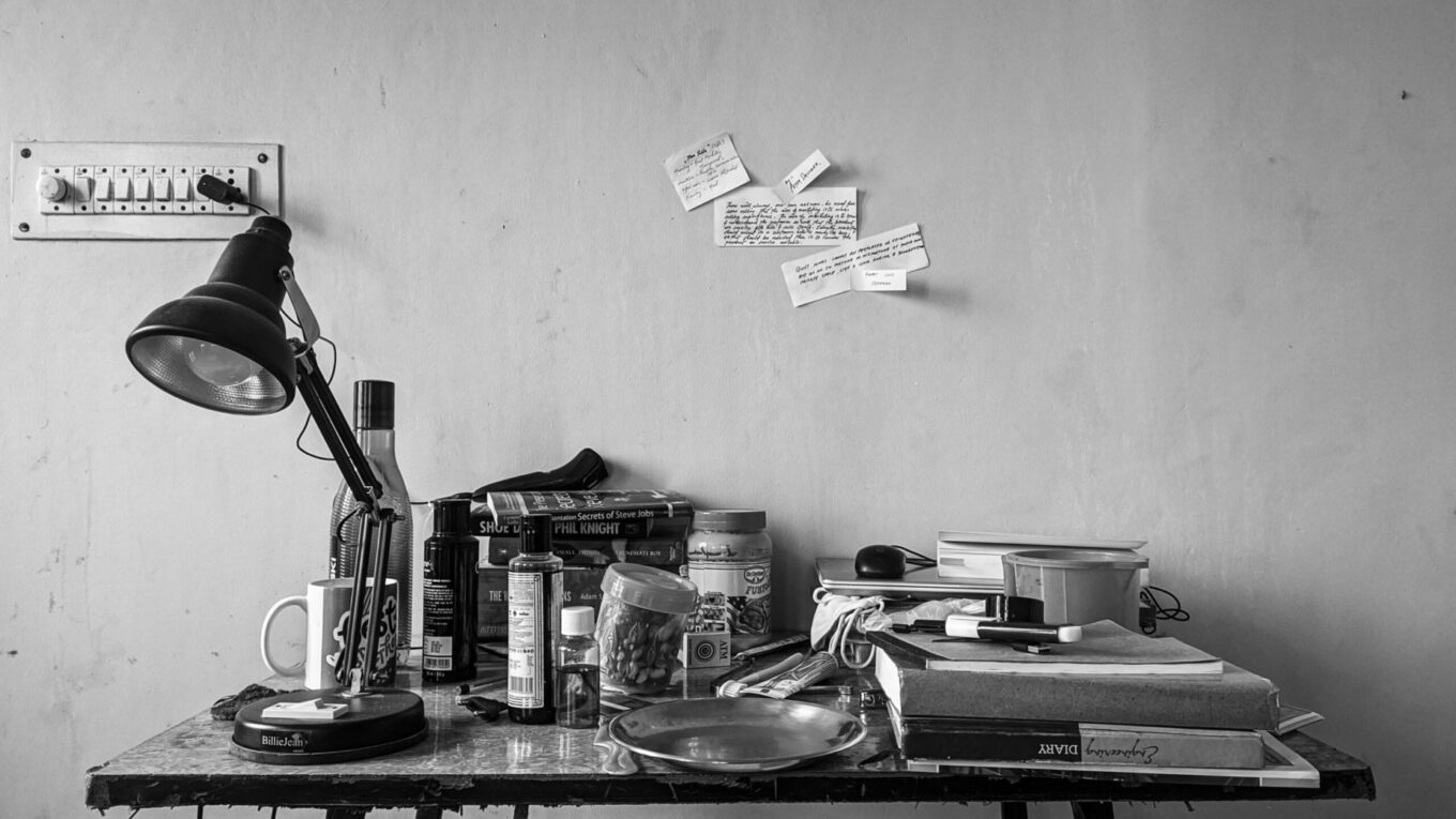 Messy table photo, in black and white, with books all over the place, post-it notes on the wall, and a broken light